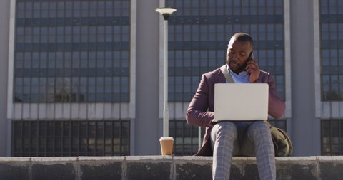 African american man in city sitting on stairs with coffee, using smartphone and laptop. digital nomad on the go, out and about in the city.