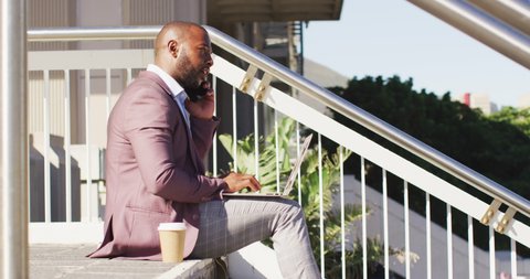 African american man in city sitting on stairs with coffee, using smartphone and laptop. digital nomad on the go, out and about in the city.
