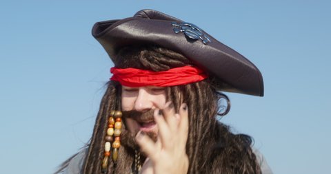 Pirate captain in cocked hat with image of skull AND crossbones above long braids rubs palms laughing against blue sky close view