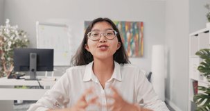 A pretty smiling Korean Asian woman with glasses sits in an office dressed in a smart white shirt, a businesswoman holds a video call, conferences, asks her boss questions over the internet