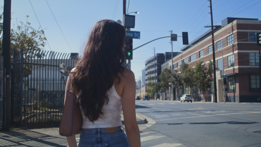 Unrecognizable dark hair woman crossing street on summer weekend. Back view young girl going crosswalk exploring city district. Stylish female tourist walking modern avenue in blue jeans on vacation.