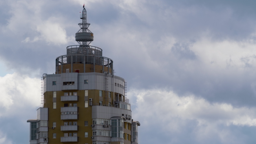 Building tower rooftop with metallic construction drone shot. Gray clouds moving. Red-black flag waving at urban multistoried architecture top on windy day. Concrete old-fashioned high-rise facade.
