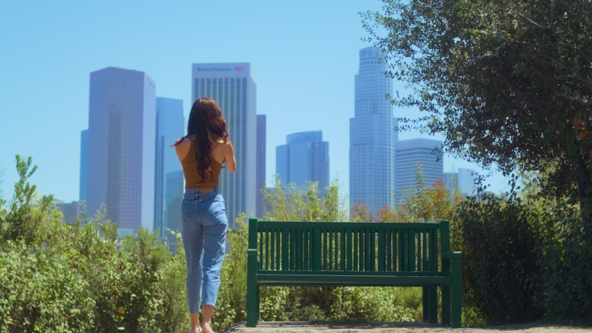 Unknown young woman standing in green park making photo skyscrapers on smartphone. Stylish girl taking picture blurred cityscape back view. Relaxed lady resting in public garden photographing far city