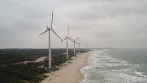 Hainan island, China - 02.05.2022: Array of onshore horizontal axis wind turbines located on a long beach coastline with high ocean swell on a cloudy day. Hainan island, China