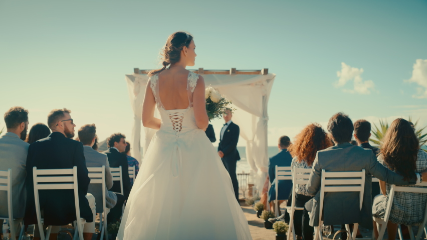 Beautiful Bride in Pure White Wedding Dress Going Down the Aisle, while Groom Waits at an Outdoors Ceremony Venue Near the Sea with Happy Multiethnic and Diverse Friends. Royalty-Free Stock Footage #1086693677