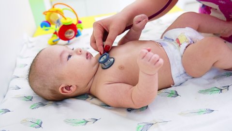Doctor’s hand puts stethoscope on baby boy’s chest. Calm beautiful kid lying peacefully on the pediatrician’s table. Close up.