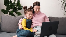 Smiling cute young mother teaching pretty little daughter to use laptop, pointing finger at screen, sitting on cozy couch at home, happy mum and adorable child girl having fun. Browsing apps concept