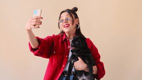 Stylish trendy woman makes a selfie online life stream video call chat on mobile phone with her pet dog, a black pug. Talking over conference recording vlog. Remote communication.