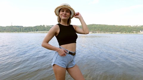 A young woman in a hat and short denim shorts walks near a river in the city. Portrait of a woman in nature