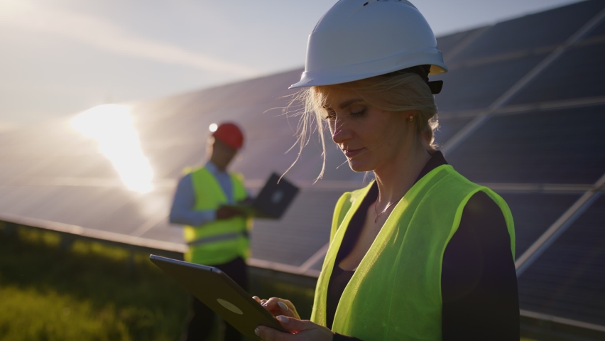 Portrait of a female solar engineer standing uses a tablet and looking at the camera, another man near the panels and entering information into a laptop. Green energy concept Royalty-Free Stock Footage #1086703382