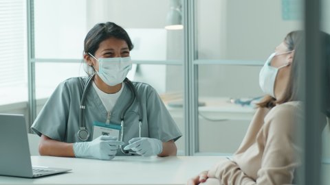 Young Hispanic midwife in medical uniform, protective mask and gloves giving consultation to pregnant woman during prenatal visit in time of coronavirus outbreak