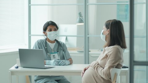 Hispanic midwife in medical uniform, protective mask and gloves speaking with Caucasian pregnant woman during prenatal checkup in time of covid-19 outbreak