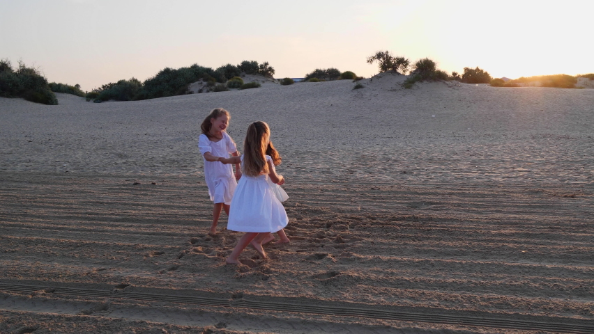 three happy sister girls in white dresses are dancing on a sandy beach at dawn in the summer and laughing merrily