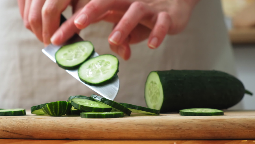 the cook cuts a spring salad of tomatoes, cucumbers, greens and mixes. Woman cutting cucumber for salad on wooden cutting board on kitchen table with cucumbers, tomatoes branch and scallion around.