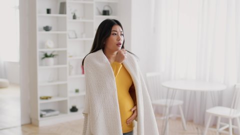 Pregnancy And Illness. Sick Pregnant Asian Woman Coughing Into Fist, Worried Expectant Mother Covered With Plaid Feeling Unwell At Home, Having Flu And Cold Symptoms, Slow Motion Footage