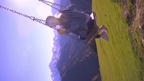 slender, beautiful blonde woman rides on large swing against background of mountains in Georgia, Svaneti heshkili. view from back. She admires view of mountains and altitude of flight. vertical video