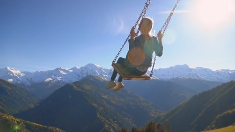 slender, beautiful blonde woman rides on large swing against background of mountains in Georgia, Svaneti heshkili. view from back. She admires view of mountains and altitude of flight. slow motion