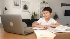 Smiling schoolboy studying on the distance from home, using trendy laptop, preteen boy doing homework, writing tasks in the notebook, watching online lessons. E-studying and homeschooling concept