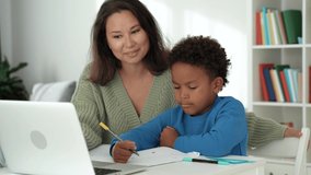 Smiling young asian mother or babysitter checking homework of smart little primary school african kid learner. Focused asian teacher educating small cute boy at private lesson at home.