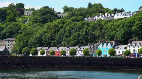 Multiple coloured houses behind the breakwater dam as a car drives along the promenade past the coloured houses on a sunny day in the town of Tobermory on the Isle of Mull. Wide shot.