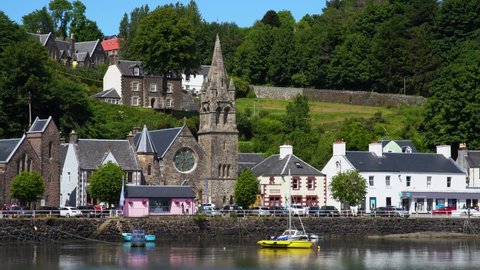 Several boatse slowly in the wind behind their anchors against the backdrop of the pretty Church of Tobermory on the Scottish island of Mull on a sunny day. Wide shot.