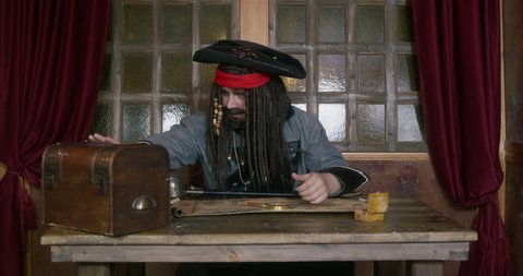 Pirate captain in cocked hat with long hair takes gold coin from treasure chest looking at map ON table against window in ship cabin