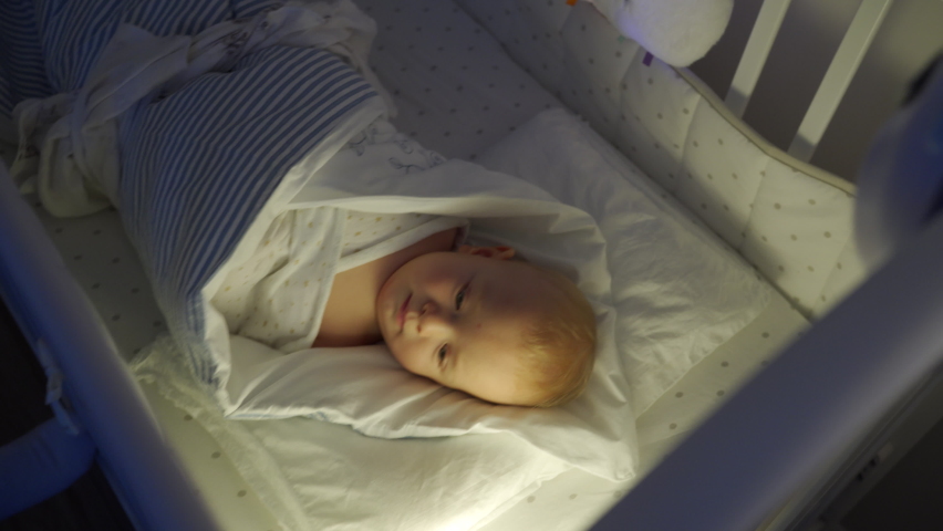 Mother rocks baby to sleep in rocking cradle in evening, swaddled 9 months baby boy in crib, face of kid in light of night lamp. High quality 4k footage Royalty-Free Stock Footage #1086752630