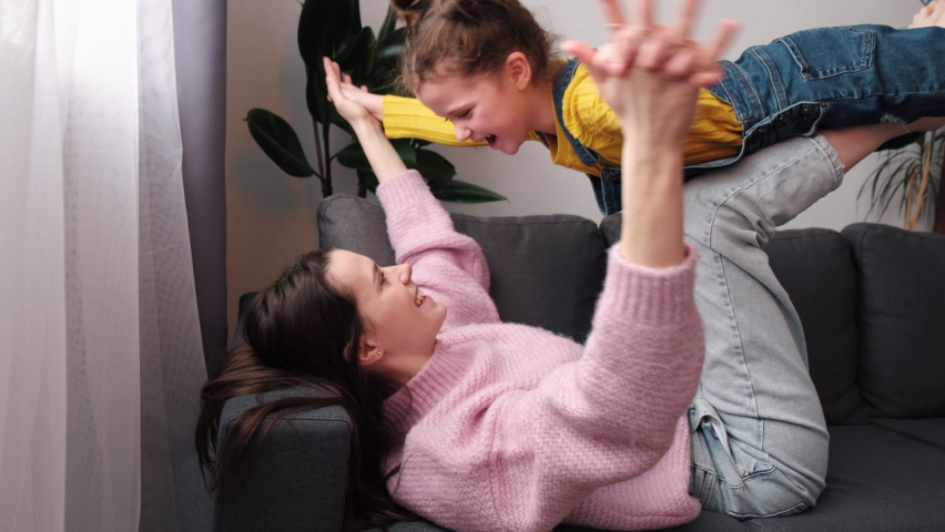 Caring young mom lying on comfy sofa on back holding little daughter in air on legs bent in knees. Carefree child girl schooler act flying doing gymnastics with mom. Play game imitate flight concept Royalty-Free Stock Footage #1086755867