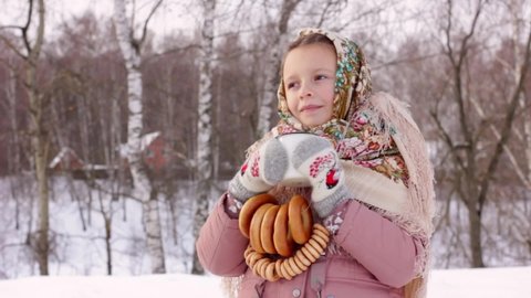 Cute girl in a traditional Russian headscarf is drinking hot tea and eating bagels on winter background. Closeup portrait of a child in folk clothes. Maslenitsa festival.