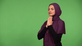 A young beautiful Muslim woman prays with her hands clasped together - green screen background