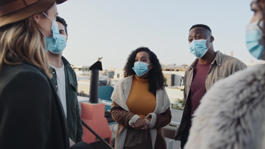 Group of adult muti-ethnic friends talking with masks on at a rooftop party at dusk.