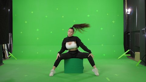 A young woman dancing on a green screen background. Attractive girl making content for social media. Female danser sits on box. Chroma key