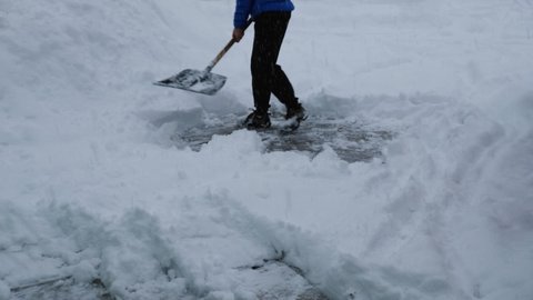 A man in a blue jacket and black pants is shoveling snow, throwing it on a large pile. Snow is falling from the sky