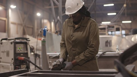 Medium shot of African American woman wearing overalls, hard hat and gloves, using grinding device on metal framework in industrial garage