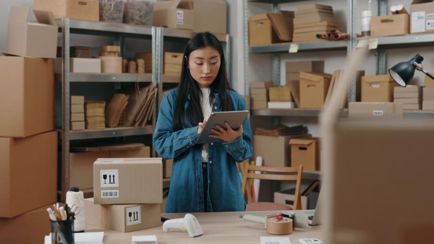 Waist up portrait view of the asian girl typing something on the tablet while checking information about parcel and preparing sending it to the client. Small business concept Royalty-Free Stock Footage #1086769511
