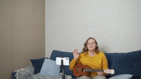 Woman conducts an online lesson and teaches students to play the ukulele. Holds guitar in hands and tells people about it via live video link. Blogging, isolation, online training