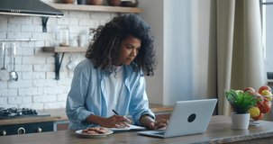 African american woman student writing notes from laptop at home. Girl distant learning, sitting at kitchen. University female studying online concentated. Education concept