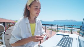 Joyful attractive woman wearing earphones, looking at laptop computer while sitting at outdoor cafe during Mediterranean vacation