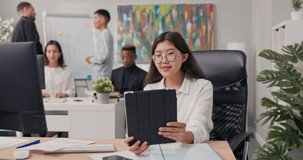 Woman in white shirt and glasses sits at desk holding tablet in hands having video conversation with boss via webcam co-workers stand at whiteboard in the background and wave hello
