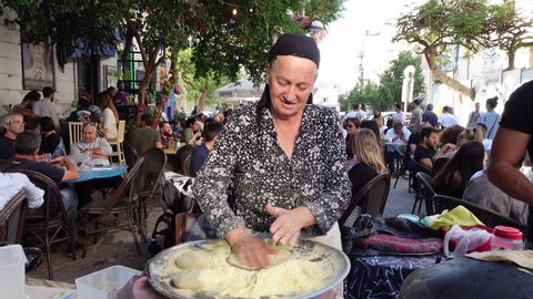 TEL AVIV, ISRAEL – NOVEMBER 29 2021: Senior Muslim woman prepares dough for flat bread at street food stall in Tel Aviv, with a busy terrace in the background. 