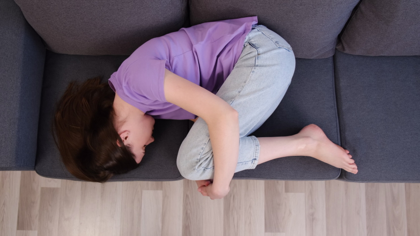 Top view of tired unhappy young woman lying on couch curled up suffers from menstrual cramps. Unplanned pregnancy and abortion, try to sleep hiding from life troubles, break up and divorce concept | Shutterstock HD Video #1086782783