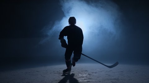 Dark silhouette of male hockey player in a uniform, helmet and with stick in his hands, skating towards the camera. An athlete skates on the ice of dark arena with backlight and smoke. Slow motion.