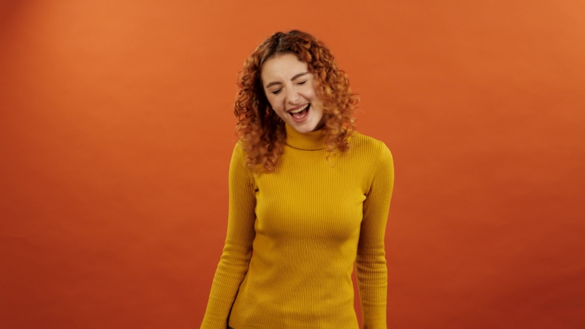 Attractive redhead caucasian girl in an orange jumper dancing and laughing to her favorite song isolated on an orange studio background. Royalty-Free Stock Footage #1086783068