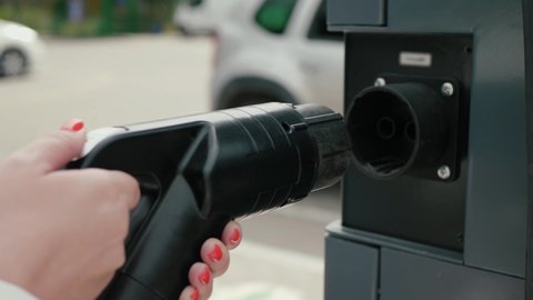 Business woman holds electric car plug against background of electric car. Female hands connect Japan type DC fast charging connector to charging station. Eco friendly alternative energy concept