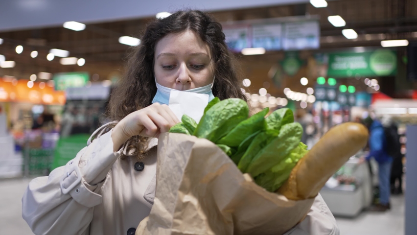 Woman shopper in a medical mask takes out a paper check from a bag of groceries in a supermarket and is very surprised at the high prices, rising inflation. Royalty-Free Stock Footage #1086793160