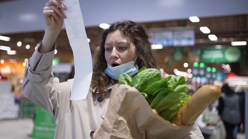 Woman shopper in a medical mask takes out a paper check from a bag of groceries in a supermarket and is very surprised at the high prices, rising inflation. | Shutterstock HD Video #1086793160