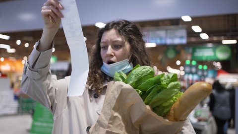Woman shopper in a medical mask takes out a paper check from a bag of groceries in a supermarket and is very surprised at the high prices, rising inflation.