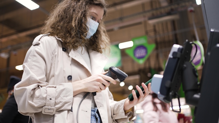 Woman in a medical mask with curly hair makes purchases at the self-service checkout in a grocery supermarket using a mobile phone Royalty-Free Stock Footage #1086793190