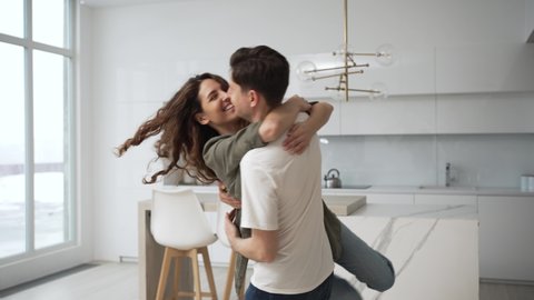 Young brunet man catches holding in arms and spins curly wife in shirt and jeans hugging tightly and laughing thankfully in kitchen closeup