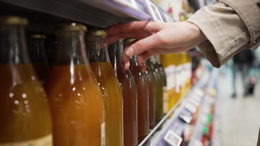 Woman's hand in a raincoat chooses fruit or vegetable healthy juice in a glass bottle in a grocery supermarket close-up